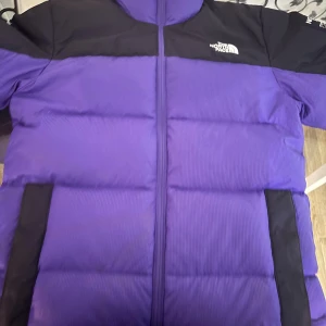 Lila pufferjacka från The North Face - Säljer en riktigt fet lila pufferjacka från The North Face med svarta detaljer på axlar och fickor. Jackan har dragkedja framtill och ett unikt färgglatt innerfoder med mönster. Perfekt för kalla dagar när du vill sticka ut.