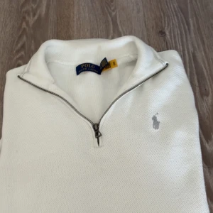 Vit half zip tröja från Polo Ralph Lauren - Snygg vit tröja från Polo Ralph Lauren med half zip och klassisk broderad logga på bröstet. Tröjan har hög krage och är tillverkad i ett stickat material som känns både stilrent och bekvämt. Perfekt för lager-på-lager och enkel att matcha.