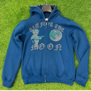 Blå hoodie från aim for the moon - Blå hoodie med huva och dragkedja framtill. Framsidan har ett stort strasstryck med texten 'AIM FOR THE MOON', en tecknad figur och en måne. Praktiska fickor och ribbade muddar vid ärmslut och nederkant. Perfekt för dig som gillar statement-plagg.