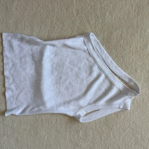 Vit one shoulder topp Pull&Bear - Säljer en vit ribbad one shoulder topp från Pull&Bear. Toppen har en asymmetrisk design med bred axelrem på ena sidan och ärmlös på andra. Materialet är mjukt och stretchigt, perfekt för en trendig och clean look.