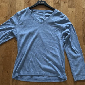 Blå långärmad tröja Tommy Hilfiger - Ljusblå långärmad tröja från Tommy Hilfiger med v-ringning och diskret logga på bröstet. Tillverkad i mjuk bomull, perfekt för en avslappnad och stilren look. Passar dig som gillar klassiska och enkla plagg.