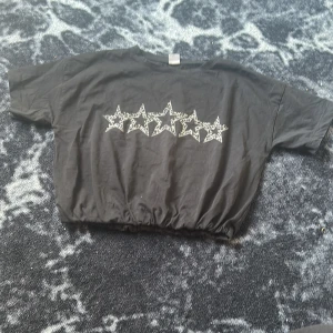 Svart croppad topp med stjärnor - Svart croppad topp i bomull med korta ärmar och resår i midjan. Framsidan har ett tryck med fem stjärnor i vitt med leopardmönster. Perfekt för dig som gillar en avslappnad men cool stil.