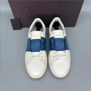 Valentino Garavani Open sneakers vit/blå - Säljer ett par snygga Valentino Garavani Open sneakers i vitt skinn med bred blå läderdetalj över vristen. Klassisk låg modell med rund tå, vita snören och diskreta nitar på hälen. Perfekta för dig som gillar stilrena och lyxiga sneakers.
