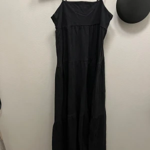 Svart långklänning med tunna axelband - En svart långklänning med tunna axelband och luftig passform. Klänningen har flera volanger och faller snyggt ner mot golvet. Perfekt för varma dagar när du vill ha något enkelt men ändå stilrent.