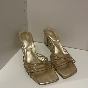 Guldiga sandaler med klack och remmar - Snygga guldiga sandaler med smala remmar och fyrkantig tå. Skorna har en elegant låg klack och öppen tå, perfekta för att lyfta din outfit 