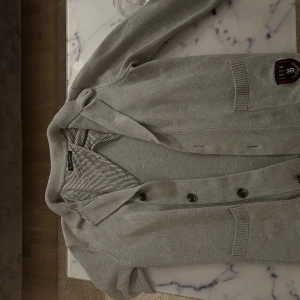Grå cardigan/kofta från Bondelid - Grå cardigan från bondelid med knappar framtill och ett broderat emblem på ena sidan. Insidan har ett randigt tyg i svart och vitt. Koftan har ribbade muddar och två fickor framtill. Perfekt för lager hösten och vintern som nu kommer! Kostar 1600 ny. Den är i storlek M men passar S också