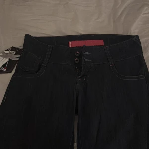 Svarta jeansbyxor från Kool Anna - Svarta jeansbyxor från Kool Anna med elastiska benslut, dubbla knappar i midjan och klassiska fickor både fram och bak. Snygga kontrastsömmar och en cool drakfluge-tag på bakfickan. Perfekta för dig som gillar streetstyle och vill ha något unikt. Dom e baggy men går in längst ner