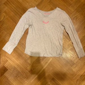 Beige långärmad tröja Zadig & Voltaire - Cool beige långärmad tröja från Zadig & Voltaire i 100% bomull. Framsidan har en liten rosa broderad detalj och baksidan har ett stort tryck med döskalle, blommor och turnétext. Avslappnad passform och rund halsringning med två små knappar.