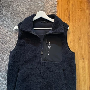 Mörkblå fleeceväst från Sail Racing - Mörkblå fleeceväst från Sail Racing med mjuk och fluffig yta. Västen har en hög krage, dragkedja framtill och en bröstficka med Sail Racing-logga. Två sidofickor med dragkedja och stilren design gör den perfekt för lager-på-lager. Passar dig som gillar sportig och clean look.