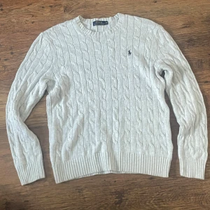 Ljusgrå kabelstickad tröja Polo Ralph Lauren - Kabelstickad ljusgrå tröja från Polo Ralph Lauren med klassisk rund halsringning och broderad marinblå logga på bröstet. Tröjan har ribbade muddar och är tillverkad i mjuk bomull. Perfekt för lager-på-lager och snygg till jeans.