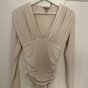 Body från H&M - Säljer en beige body från H&M med djup v-ringning och långa ärmar. bodyn har snygga rynkade detaljer vid axlar och midja som ger en cool siluett. Perfekt att styla med jeans eller kjol för en trendig look.