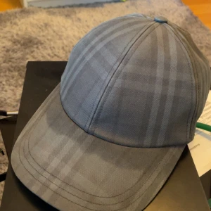 Grå rutig Burberry keps - Snygg grå keps från Burberry med diskret rutigt mönster och broderad logga baktill. Justerbar rem för perfekt passform. Tillverkad i bomull som känns skön och lätt på huvudet. Perfekt för dig som vill ha en stilren och klassisk look. Den är i jätte bra skick förutom solblekning. Det bör gå att fixa men orkar verkligen inte