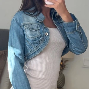 Croppad jeansjacka med knappar - Säljer en croppad ljusblå jeansjacka med coola detaljer som fickor med lock och silvriga knappar framtill. Jackan har lång ärm och klassisk krage, perfekt att slänga över en topp för en chill look.