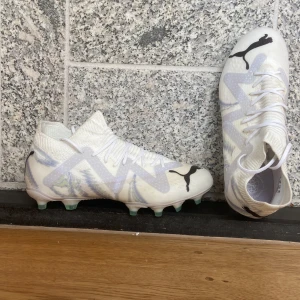 Helt oanvänd. Puma future 6 Elite FG/AG Brilliance Puma White Limited edition  - Säljer ett par vita Puma fotbollsskor med svart logga och coolt mönster i ljusa toner. Skorna har snörning, stickad strumpliknande krage och platt sula med gröna dobbar. Perfekta för dig som vill sticka ut på planen. Skorna är Elite alltså bästa modellen.