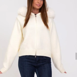 Vit stickad cardigan jacka med huva S/M - Mysig vit stickad cardigan jacka med dragkedja och fluffig huva med pälsdetalj. Jackan har ribbade muddar och en avslappnad passform, perfekt för kyliga dagar. Enkel och stilren design som passar till jeans eller kjol.