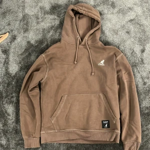 Brun hoodie från Pull&Bear x Kangol - Säljer en brun hoodie från Pull&Bear x Kangol med klassisk känguru-ficka och snörning i huvan. Tröjan har ribbade muddar och en vit Kangol-logga på bröstet. Mjuk insida och relaxed passform, perfekt för chill dagar.