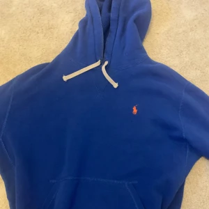 Blå hoodie från Polo Ralph Lauren - Snygg blå hoodie från Polo Ralph Lauren med klassisk röd logga på bröstet. Tröjan har huva med vit snörning, magficka och långa ärmar. Mjuk insida och ribbade muddar vid ärmslut och nederkant. Perfekt för chill dagar och streetwear. Jag är 185 cm och den sitter lite kort på mig👍🏻