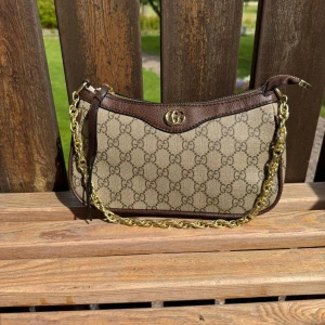 Gucci axelväska med GG-mönster - Snygg axelväska från Gucci i beige canvas med klassiskt GG-mönster och bruna skinn-detaljer. Väskan har guldfärgad kedja, dragkedja upptill och logga framtill. Perfekt storlek för det viktigaste och en riktig statement piece.