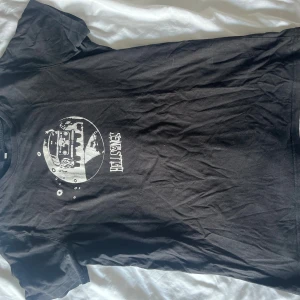 Svart Hellsongs t-shirt från Jem Collective - Svart t-shirt från Jem Collective med vit Hellsongs-tryck framtill. Trycket visar en bil med blommor och flames i en cirkel, samt texten Hellsongs. Klassisk passform och rund hals. Perfekt för dig som gillar bandmerch och coola prints.