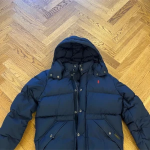 Mörkblå pufferjacka från Polo Ralph Lauren - Säljer en mörkblå pufferjacka från Polo Ralph Lauren med huva och snygga detaljer som röda loggan på bröstet. Jackan har dragkedja och knappar framtill, två fickor med lock och tryckknappar. Perfekt för kalla dagar och riktigt chill stil. Pris kan gå ner vid snabb affär!!