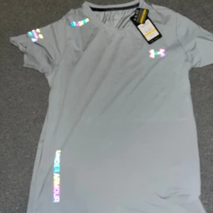 Grå Under Armour t-shirt M - Snygg grå t-shirt från Under Armour med holografiska loggor på bröstet, axeln och vid sidan. Tillverkad i lätt och ventilerande polyester, perfekt för träning. Klassisk passform med rund hals och korta ärmar. Modell: Threadborne.
