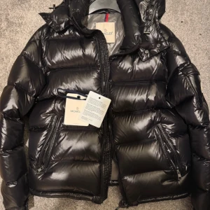 Svart pufferjacka från Moncler - Säljer en svart, glansig pufferjacka från Moncler med huva och dragkedja framtill.                                   Moncler dustbag, bild på kvittot, Moncler tagg och så även Moncler nfc kod kommer att följa med. Vid funderingar så är det bara kontakta 👍