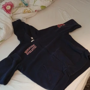 Mörkblå hoodie från Nonation Athletic - Mörkblå hoodie med dragkedja från Nonation Athletic. Tryck med texten 'Nonation Athletic' i vitt och rött på bröstet och en stor röd 3:a på ena ärmen. Klassisk känguruficka framtill och huva. Perfekt för en sportig och avslappnad stil.