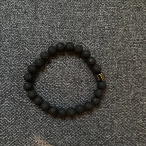 Svart armband med lavapärlor från Bvaldi - Stilrent armband med svarta lavapärlor och en liten guldfärgad detalj från Bvaldi. Elastiskt band gör det enkelt att ta på och av. Perfekt accessoar för en cool och minimalistisk look.