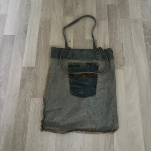 Jeansväska från Nudie Jeans med ficka - Unik shoppingväska gjord av återvunna jeans från Nudie Jeans. Väskan har en cool blå jeansfärg med kontrastsömmar och en stor ficka framtill med Nudie Jeans-logga. Långa axelband och metallnitar upptill ger extra detaljer. Perfekt för dig som gillar street och vintage vibes.