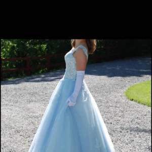 Ljusblå långklänning med spetsdetaljer - ljusblå långklänning med spetsapplikationer och blommiga broderier på kjolen. Klänningen har v-ringad urringning, breda axelband och är i storlek 32, men passar även 34. jag är 160 cm men passar upp till 172 cm utan att den blir för kort. hör gärna av dig om du är intresserad även om det bara är för att testa klänningen!