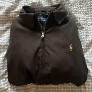 Svart vindjacka från Polo Ralph Lauren - Snygg svart vindjacka från Polo Ralph Lauren med klassisk krage, dragkedja framtill och broderad logga på bröstet. Jackan har två snedställda fickor och elastisk! Använd ett fåtal gånger! Pris går att diskutera, kontakta vid frågor! 
