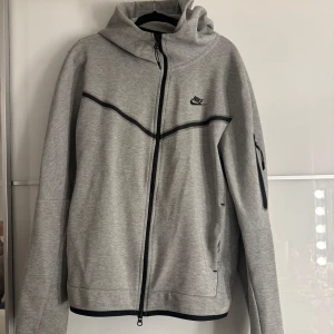 Nike tech hoodie - Säljer en grå hoodie från Nike med hel dragkedja och svart logga på bröstet. Tröjan har huva, långa ärmar och en praktisk ficka med dragkedja på ena ärmen. Perfekt för chill eller träning.