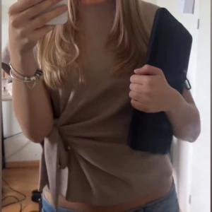 Beige topp med knyt från Zara - Säljer en beige topp från Zara med korta ärmar och rund halsringning. Toppen har en snygg knytdetalj i sidan som ger en cool twist till looken. Materialet är mjukt och lätt ribbat, perfekt till jeans eller kjol.