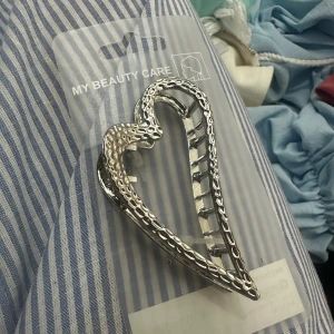 Silverfärgad hjärtformad hårklämma - Säljer en snygg hårklämma i silverfärg från My Beauty Care. Klämman har en unik hjärtformad design med ett coolt mönster längs kanterna som ger den extra edge. Perfekt accessoar för att lyfta din frisyr och ge en trendig touch.