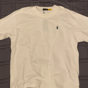 Vit t-shirt från Polo Ralph Lauren (prisförslag) - Vit t-shirt från Polo Ralph Lauren med klassisk rund hals och korta ärmar. På bröstet finns den ikoniska lilla broderade Polo-loggan i marinblått. T-shirten är gjord i mjuk bomull och har en clean, stilren look som funkar till allt.