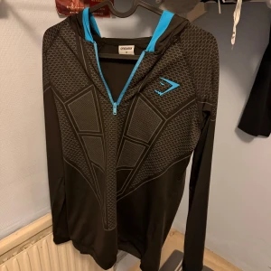 Onyx v1 hoodie M - Svart hoodie från Gymshark med coolt geometriskt mönster och blå detaljer vid dragkedja och huva. Långärmad och tillverkad i mjukt syntetmaterial, perfekt för träning. Gymshark-logga i blått på bröstet. Snygg och sportig stil!