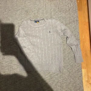 Grå stickad tröja Polo Ralph Lauren - Snygg grå stickad tröja från Polo Ralph Lauren med klassisk kabelstickad design och broderad marinblå logga på bröstet. Tröjan har rund hals och långa ärmar med ribbade muddar. Perfekt för dig som gillar stilrena och tidlösa plagg.