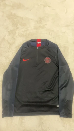 PSG långärmad träningströja Nike står M men passar S  - Snygg långärmad PSG träningströja från Nike i mörkblått med röd Nike-logga och PSG-emblem på bröstet. Tröjan har halv dragkedja och är tillverkad i ett lätt, tekniskt material som andas. Perfekt för fotbollsträning eller att representera ditt lag.