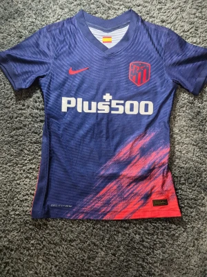 Atlético Madrid blå matchtröja Nike - Säljer en officiell Atlético Madrid fotbollströja från Nike i mörkblått med röda detaljer och klubbmärke på bröstet. Tröjan har korta ärmar, V-ringad krage och tryckt sponsor Plus500 på framsidan. Materialet är lätt och andas, perfekt för träning eller match.