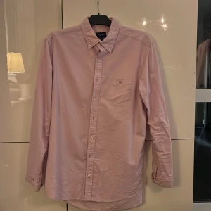 Ljusrosa Oxfordskjorta från GANT - Snygg ljusrosa Oxfordskjorta från GANT i storlek L. Skjortan har klassisk button-down krage, bröstficka med broderad logga och vita knappar. Regular fit och tillverkad i mjuk bomull. Perfekt för dig som gillar stilrena och tidlösa plagg.