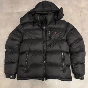 Svart pufferjacka från Polo Ralph Lauren - Svart pufferjacka från Polo Ralph Lauren med huva och flera dragkedjeförsedda fickor. Jackan har det klassiska röda Polo-logot broderat på bröstet och är tillverkad i syntetmaterial. Perfekt för kalla dagar med sin quiltade design och snygga, sportiga look. Priset kan diskuteras om du verkligen är intresserad det är 12-14 år