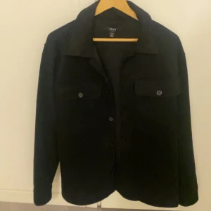 Svart overshirt  - Svart overshirt med klassisk krage och knäppning framtill. Två bröstfickor med knappstängning ger en clean och stilren look. Perfekt att bära som lager över t-shirt eller hoodie. Långärmad och i mjukt material.