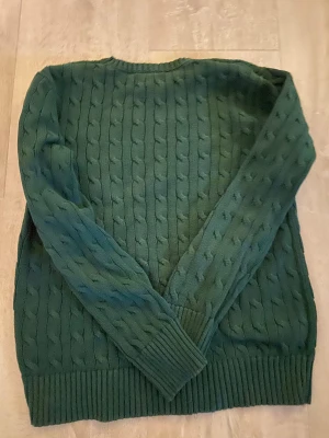 Grön kabelstickad tröja Polo Ralph Lauren - Mörkgrön kabelstickad tröja från Polo Ralph Lauren i storlek S. Tröjan har rund hals, långa ärmar och breda ribbade muddar vid ärmslut och nederkant. Perfekt för dig som gillar klassisk stil med snygga detaljer.