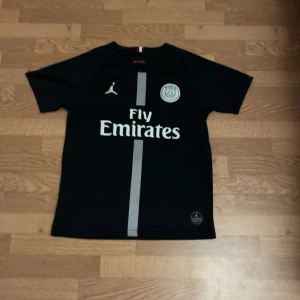 PSG Neymar Jr svart fotbollströja - Svart PSG-matchtröja med Neymar Jr och nummer 10 på ryggen. Framsidan har Fly Emirates-tryck, PSG-logga och Jordan-logga. Tröjan har en vit vertikal rand i mitten och är tillverkad i lätt, ventilerande material. Kortärmad och sportig design.