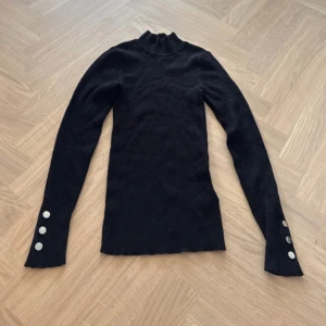 Svart ribbad polotröja från Amisu - Svart polotröja från Amisu i ribbad stickad kvalitet. Tröjan har lång ärm med tre silvriga knappar vid ärmslutet och en tight passform. Perfekt att styla med jeans eller kjol för en clean look.