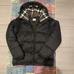 Burberry - Pufferjacka  - Svart pufferjacka från Burberry med klassiskt rutigt foder i huvan. Jackan har dragkedja och knappar framtill, justerbar huva och ribbade muddar. Yttermaterialet är i bomull och jackan har en riktigt clean och tidlös look. Har sytt ihop armarna med jätte lite sömmar, men det går att sprätta upp enkelt, så de blir till en väst. Jackan är använd fåtal gånger, och är i storlek L men passar även M. Tveka inte med att skriva🐻