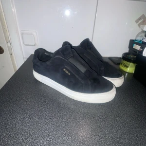 Axel Arigato skor  - Säljer ett par svarta sneakers från Axel Arigato med stilren design och vit platt sula. Skorna är tillverkade i mjuk mocka och har elastiska band istället för klassisk snörning. Diskret Arigato-logga i guld på sidan. Perfekta för dig som gillar minimalistisk stil. Säljs 1,8 föraren till kvarn som vita, strolek 42