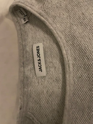 Ljusgrå stickad tröja Jack & Jones XS - Snygg ljusgrå stickad tröja från Jack & Jones i storlek XS. Tröjan har en enkel design med rund hals och korta ärmar, perfekt för dig som gillar stilrena och bekväma plagg. Materialet är mjukt och har en diskret struktur som ger en cool känsla. Priset kan diskuteras