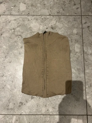 Beige stickad kofta med dragkedja - Snygg beige stickad kofta med ribbad hög krage och hel dragkedja framtill. Perfekt för lager-på-lager och chill stil. Långärmad och med mjukt material som känns skönt mot huden. Enkel att matcha med jeans eller sweatpants.