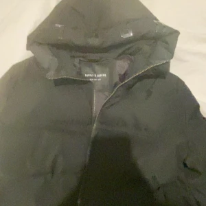 Svart pufferjacka Supply & Demand - Svart pufferjacka från Supply & Demand med huva och dragkedja. Jackan är vadderad och har en klassisk pufferlook som håller dig varm under kalla dagar. Perfekt för dig som gillar streetstyle och vill ha en snygg och enkel jacka till vintern.